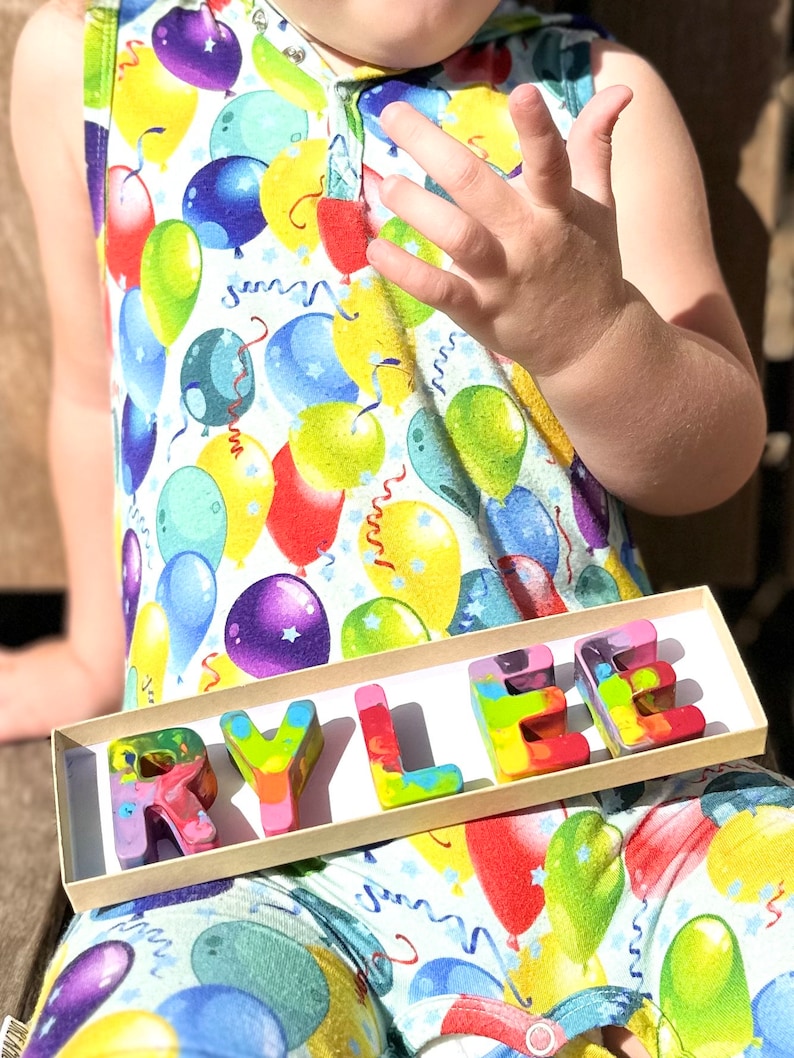 May include: A child wearing a sleeveless jumpsuit with a colorful balloon print holds a tray of rainbow-colored crayons spelling out the name "RYLEE". The crayons are in a rectangular box.