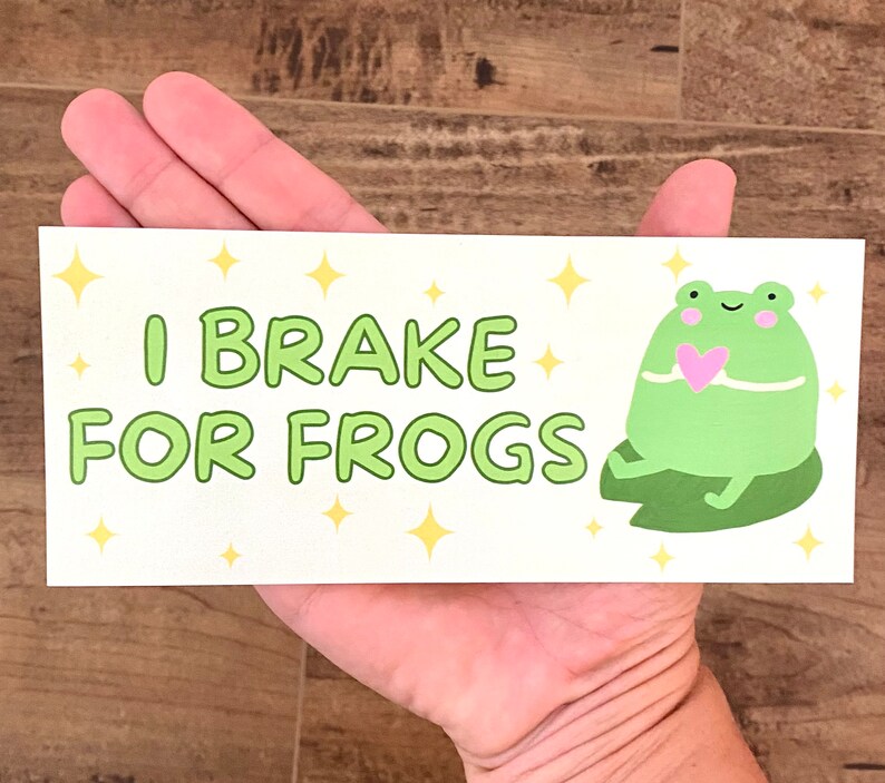 May include: A white sticker with green text that reads "I Brake For Frogs". A green frog with a pink heart is sitting on a green lily pad. The sticker has a white background with yellow stars.
