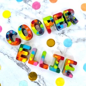 May include: Colorful, handmade crayon letters spelling "COOPER" and "ELLIE" on a marble surface, surrounded by confetti. The letters are a mix of vibrant colors, including red, yellow, green, and blue, creating a playful and artistic look.