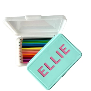 May include: A turquoise pencil case with a pink inscription that reads "ELLIE". The case is open and contains a set of colored pencils.