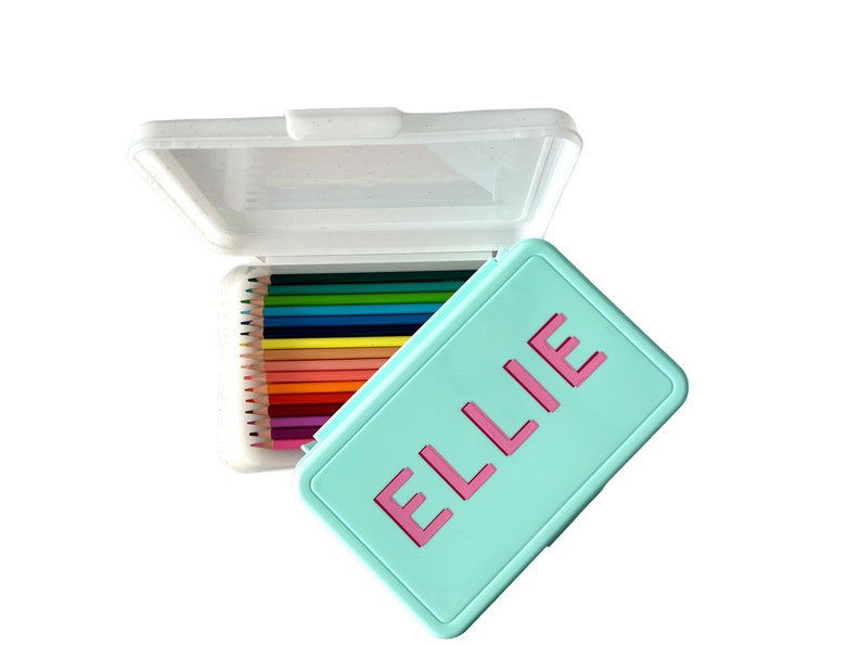 May include: A turquoise pencil case with a pink inscription that reads "ELLIE". The case is open and contains a set of colored pencils.