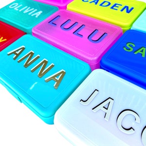 May include: A set of colorful rectangular lunch boxes with names printed on them. The names include Olivia, Caden, Lulu, Sammy, Anna, Sam, and Jacob.