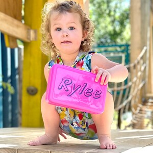 May include: A young girl with curly blonde hair is squatting on a wooden platform. She is holding a pink lunchbox with the name "Rylee" written in purple letters.