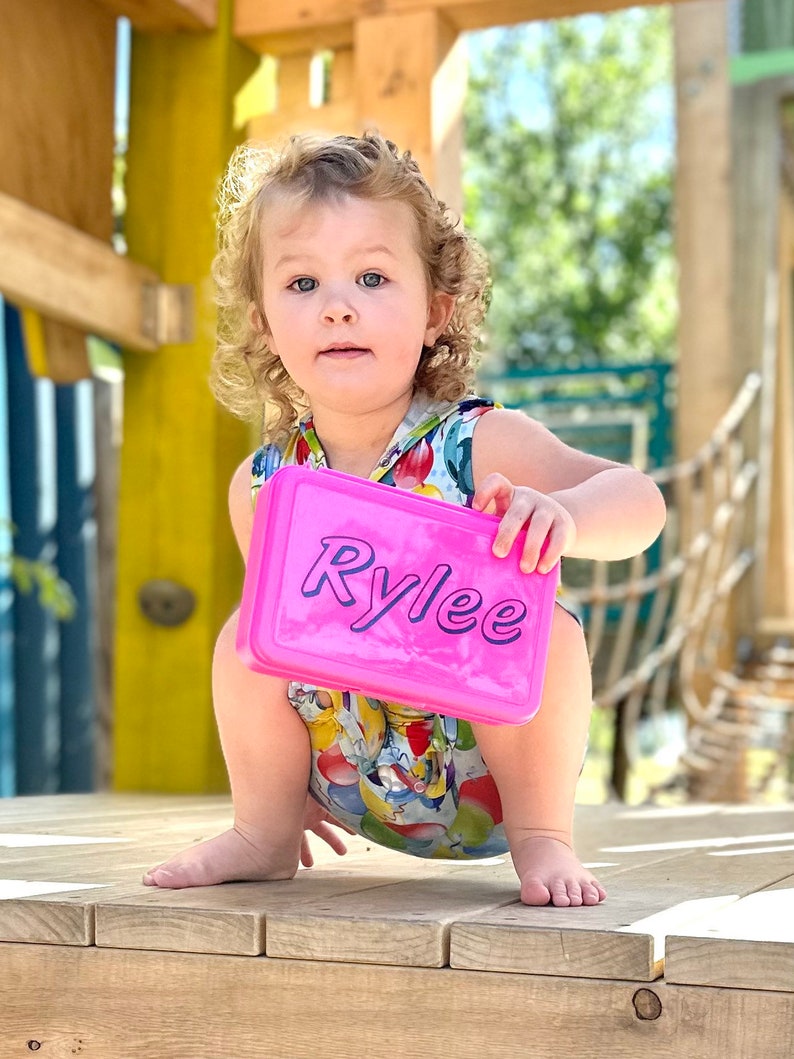 May include: A young girl with curly blonde hair is squatting on a wooden platform. She is holding a pink lunchbox with the name "Rylee" written in purple letters.