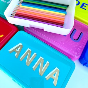 May include: A set of colorful pencil cases with glitter. One case is turquoise blue with the name "ANNA" in gold letters. Another case is pink with glitter and the letter "U". A third case is yellow with the letter "DE". The fourth case is white with a row of colored pencils inside.