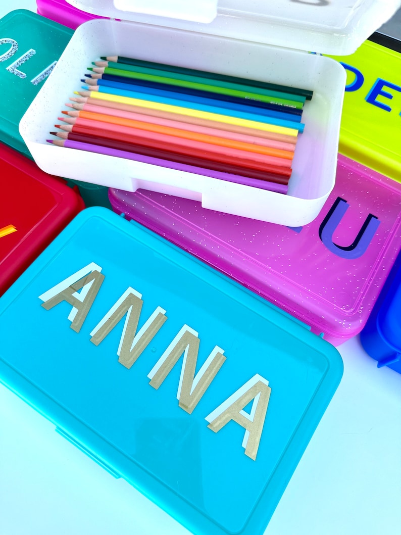 May include: A set of colorful pencil cases with glitter. One case is turquoise blue with the name "ANNA" in gold letters. Another case is pink with glitter and the letter "U". A third case is yellow with the letter "DE". The fourth case is white with a row of colored pencils inside.
