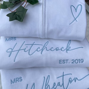 Puede incluir: Tres sudaderas blancas con cremallera y bordado en azul claro. La sudadera superior tiene un contorno de corazón. La sudadera central muestra el texto "MRS Hitchcock EST. 2019". La sudadera inferior muestra el texto "MRS Wheaton EST. 202".