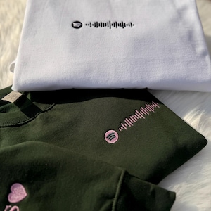 May include: White and dark green sweatshirts with embroidered Spotify codes. The white sweatshirt has a black code, while the dark green sweatshirts feature pink codes. One dark green sweatshirt has a pink heart and the letter "S".