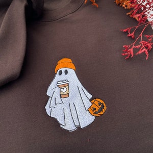 May include: A brown sweatshirt featuring an embroidered design of a ghost wearing an orange hat and holding a coffee cup. The ghost is carrying a small orange pumpkin.