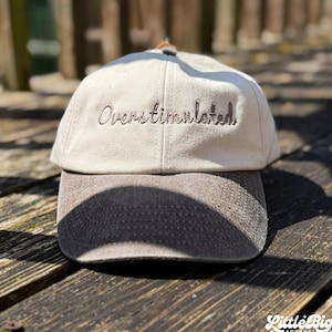Könnte beinhalten: Eine zweifarbige Baseballkappe mit cremeweißer Krone und dunkelgrauem Schirm. Das Wort Overstimulated ist in dunklem Garn auf die Vorderseite der Kappe gestickt. Das Design umfasst eine strukturierte Vorderseite und einen gebogenen Schirm.