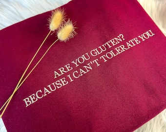 May include: A deep maroon t-shirt featuring white embroidered text that reads, ARE YOU GLUTEN? BECAUSE I CAN'T TOLERATE YOU. Two dried, light tan, fluffy botanical stems are placed diagonally across the upper left corner of the fabric.