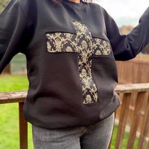 Puede incluir: Sudadera negra con un diseño de cruz grande. La cruz está hecha de una tela dorada y negra estampada. La sudadera tiene cuello redondo y mangas largas. Un estilo casual y cómodo.