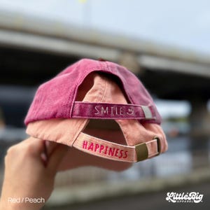 Puede incluir: Una gorra de béisbol roja y melocotón con la palabra "SMILE" bordada en la correa trasera. La correa inferior tiene la palabra "HAPPINESS" bordada en rojo. La gorra está hecha de un material suave.