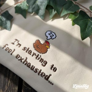 Puede incluir: Sudadera de color crema con texto bordado y un gr&aacute;fico de pollo pixelado. El texto dice "I'm starting to feel exhausted..." Una burbuja de pensamiento sobre el pollo muestra el n&uacute;mero 22. La sudadera tiene un peque&ntilde;o logotipo de "Little Big Apparel".