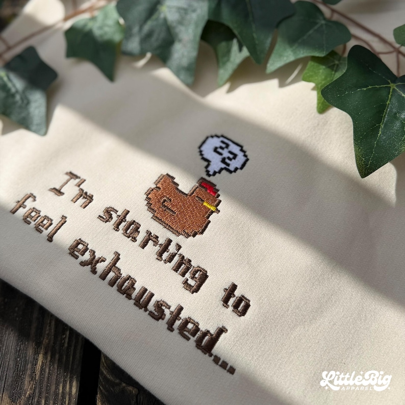 Puede incluir: Sudadera de color crema con texto bordado y un gr&aacute;fico de pollo pixelado. El texto dice "I'm starting to feel exhausted..." Una burbuja de pensamiento sobre el pollo muestra el n&uacute;mero 22. La sudadera tiene un peque&ntilde;o logotipo de "Little Big Apparel".