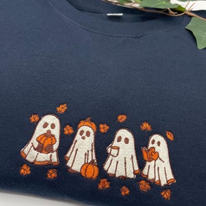 May include: Navy blue sweatshirt with an embroidered design of four cartoon ghosts holding pumpkins, a coffee cup, and a book. The ghosts are surrounded by orange autumn leaves, ideal for Halloween or autumn.