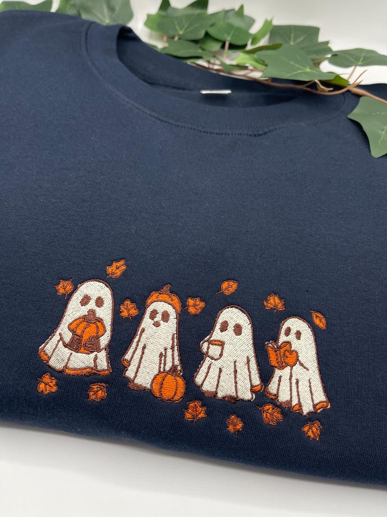 May include: Navy blue sweatshirt with an embroidered design of four cartoon ghosts holding pumpkins, a coffee cup, and a book. The ghosts are surrounded by orange autumn leaves, ideal for Halloween or autumn.
