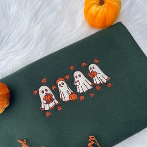 May include: Dark green sweatshirt with an embroidered design of four cartoon ghosts holding pumpkins. The ghosts are white with orange accents, and the pumpkins are orange. Scattered orange leaves surround the ghosts. Two small orange pumpkins are in the background.