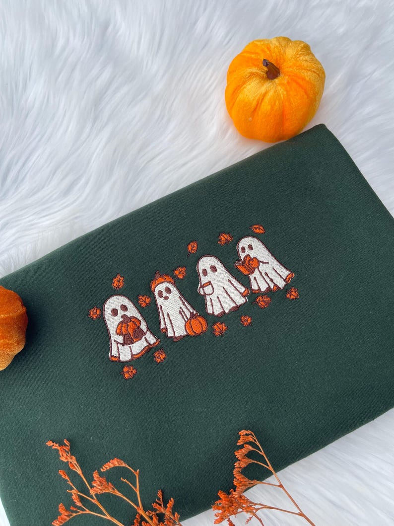 May include: Dark green sweatshirt with an embroidered design of four cartoon ghosts holding pumpkins. The ghosts are white with orange accents, and the pumpkins are orange. Scattered orange leaves surround the ghosts. Two small orange pumpkins are in the background.