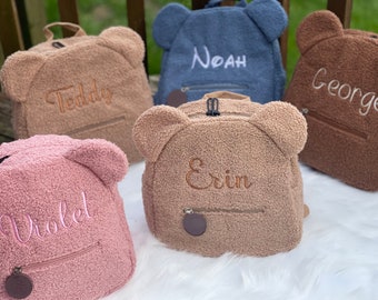 May include: Five plush teddy bear backpacks with embroidered names: Teddy, Noah, George, Erin, and Violet. The backpacks are in shades of brown, blue, and pink.