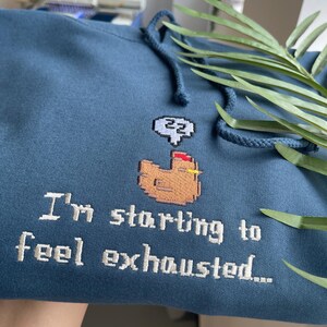 Puede incluir: Sudadera azul con capucha y cord&oacute;n ajustable con dise&ntilde;o bordado. El dise&ntilde;o incluye un pollo pixelado con una burbuja de pensamiento y el texto "I'm starting to feel exhausted..."