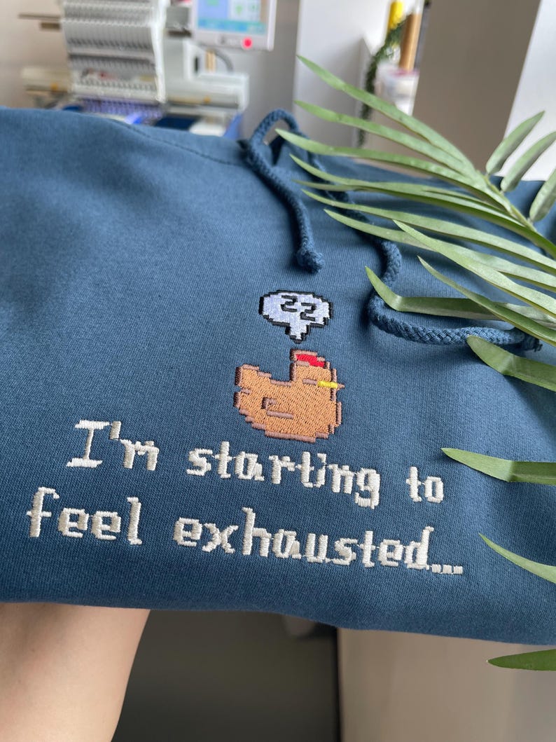 Puede incluir: Sudadera azul con capucha y cord&oacute;n ajustable con dise&ntilde;o bordado. El dise&ntilde;o incluye un pollo pixelado con una burbuja de pensamiento y el texto "I'm starting to feel exhausted..."