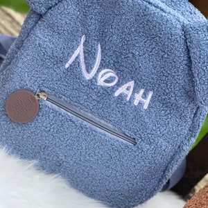 K&ouml;nnte beinhalten: Ein blauer Pl&uuml;sch-Rucksack mit Rei&szlig;verschluss und einem B&auml;ren-Rei&szlig;verschlussanh&auml;nger. Der Rucksack hat den Namen "Noah" in wei&szlig; auf der Vorderseite gestickt.