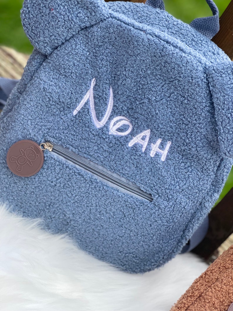 K&ouml;nnte beinhalten: Ein blauer Pl&uuml;sch-Rucksack mit Rei&szlig;verschluss und einem B&auml;ren-Rei&szlig;verschlussanh&auml;nger. Der Rucksack hat den Namen "Noah" in wei&szlig; auf der Vorderseite gestickt.