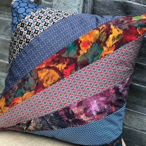 May include: A square cushion with a patchwork design made from different patterned fabrics. The cushion is made from a variety of blue, red, and orange fabrics with geometric and floral patterns.