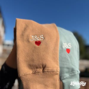 Op de afbeelding: Close-up van twee sweatshirts, één bruin en één blauwgroen, met geborduurde initialen "J&S" en een rood hart. Het bruine sweatshirt staat op de voorgrond, met de blauwgroene erachter. Het logo "Little Big Apparel" is rechtsonder zichtbaar.