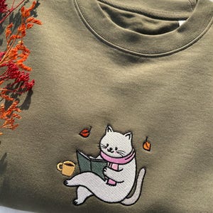 May include: A green sweatshirt with an embroidered design of a white cat wearing a pink scarf and reading a book. The cat is sitting with a cup of coffee next to it. There are also some leaves falling around the cat.