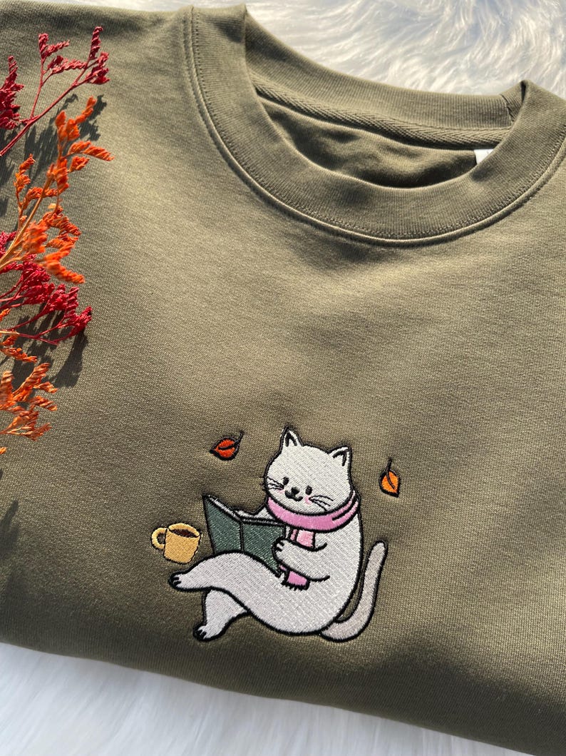 May include: A green sweatshirt with an embroidered design of a white cat wearing a pink scarf and reading a book. The cat is sitting with a cup of coffee next to it. There are also some leaves falling around the cat.