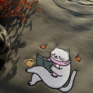 May include: A green sweatshirt with an embroidered design of a white cat reading a book while wearing a pink scarf. The cat is sitting on a green surface with a cup of coffee and leaves scattered around it.