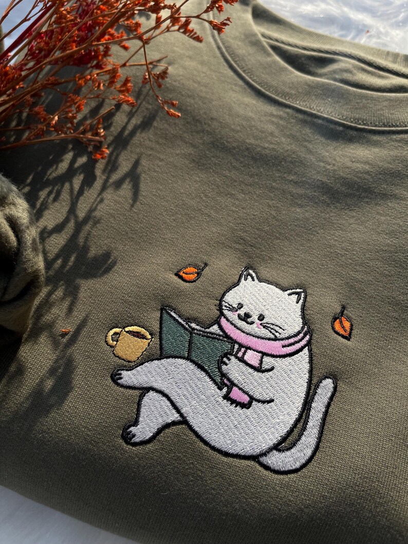 May include: A green sweatshirt with an embroidered design of a white cat reading a book while wearing a pink scarf. The cat is sitting on a green surface with a cup of coffee and leaves scattered around it.