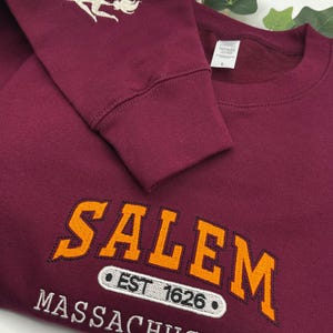 Peut inclure: Sweat-shirt bordeaux avec le mot "SALEM" en orange, soulign&eacute; de noir, et le texte "MASSACHUSETTS" en dessous. La manche pr&eacute;sente une sorci&egrave;re argent&eacute;e sur un balai. Le texte "EST 1626" est &eacute;galement visible.