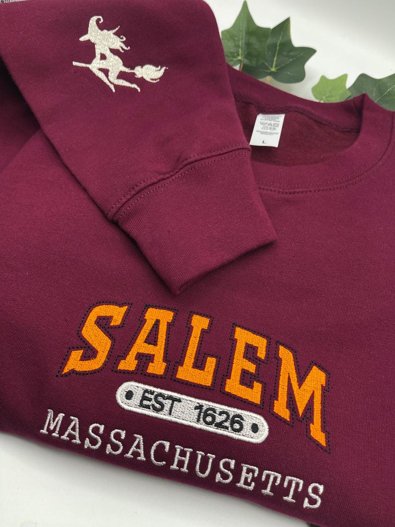 Peut inclure: Sweat-shirt bordeaux avec le mot "SALEM" en orange, soulign&eacute; de noir, et le texte "MASSACHUSETTS" en dessous. La manche pr&eacute;sente une sorci&egrave;re argent&eacute;e sur un balai. Le texte "EST 1626" est &eacute;galement visible.