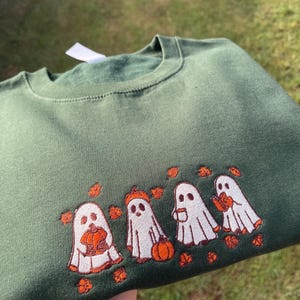 May include: Dark green sweatshirt featuring an embroidered design of four white ghosts with orange details. Each ghost holds a pumpkin, a pumpkin hat, a cup, and a leaf. The design is surrounded by orange leaves.