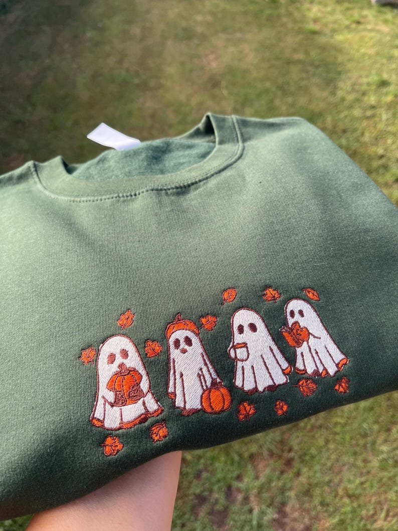 May include: Dark green sweatshirt featuring an embroidered design of four white ghosts with orange details. Each ghost holds a pumpkin, a pumpkin hat, a cup, and a leaf. The design is surrounded by orange leaves.