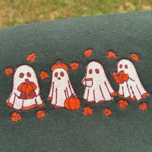 May include: Dark green sweatshirt with an embroidered design of four white ghosts with orange detailing. Each ghost holds a pumpkin, wears a pumpkin hat, holds a mug, or reads a book. Orange leaves are scattered around.