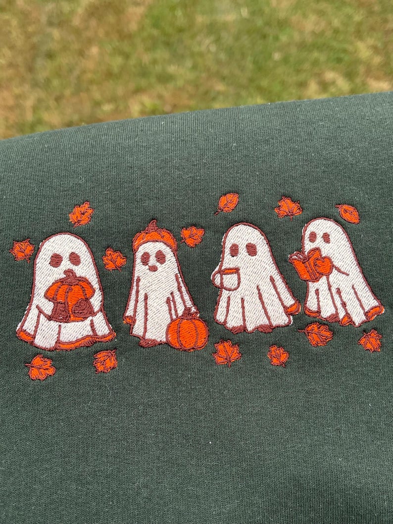 May include: Dark green sweatshirt with an embroidered design of four white ghosts with orange detailing. Each ghost holds a pumpkin, wears a pumpkin hat, holds a mug, or reads a book. Orange leaves are scattered around.