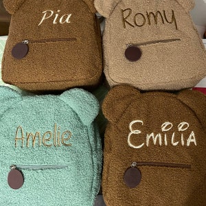 K&ouml;nnte beinhalten: Vier braune und gr&uuml;ne Pl&uuml;sch-Rucksack-Teddyb&auml;ren mit Rei&szlig;verschlusstaschen und B&auml;renohren. Die Rucks&auml;cke sind mit den Namen Pia, Romy, Amelie und Emilia personalisiert.