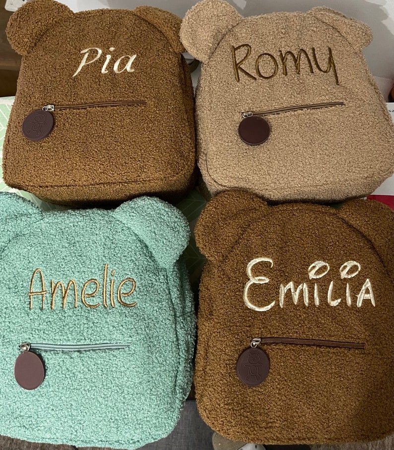 K&ouml;nnte beinhalten: Vier braune und gr&uuml;ne Pl&uuml;sch-Rucksack-Teddyb&auml;ren mit Rei&szlig;verschlusstaschen und B&auml;renohren. Die Rucks&auml;cke sind mit den Namen Pia, Romy, Amelie und Emilia personalisiert.
