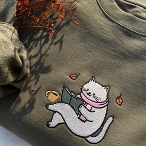May include: A green sweatshirt with an embroidered design of a cat reading a book and holding a cup of coffee. The cat is wearing a pink scarf and there are falling leaves around it.