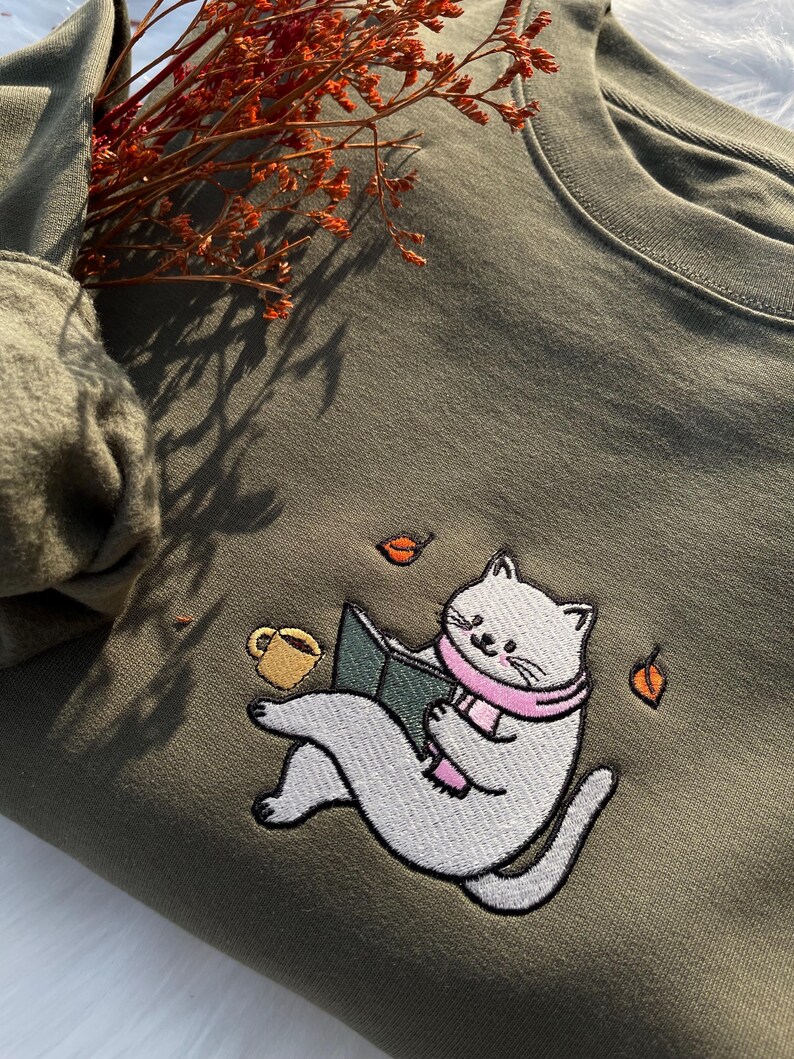 May include: A green sweatshirt with an embroidered design of a cat reading a book and holding a cup of coffee. The cat is wearing a pink scarf and there are falling leaves around it.