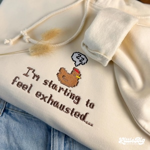 Puede incluir: Sudadera con capucha color crema con cord&oacute;n y un gr&aacute;fico de pollo pixelado. El texto "I'm starting to feel exhausted..." est&aacute; bordado debajo. La sudadera est&aacute; doblada sobre mezclilla azul.