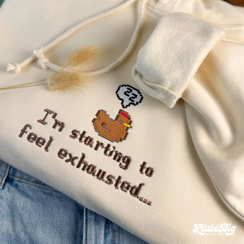 Puede incluir: Sudadera con capucha color crema con cord&oacute;n y un gr&aacute;fico de pollo pixelado. El texto "I'm starting to feel exhausted..." est&aacute; bordado debajo. La sudadera est&aacute; doblada sobre mezclilla azul.