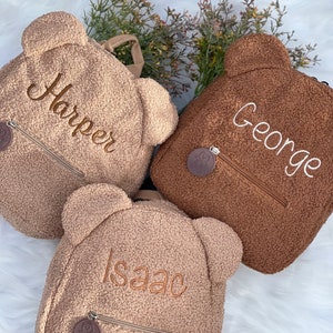K&ouml;nnte beinhalten: Drei braune Teddyb&auml;r-Rucks&auml;cke mit personalisierten Namen: Harper, George und Isaac. Jeder Rucksack hat einen kleinen braunen B&auml;ren-Anh&auml;nger am Rei&szlig;verschluss.