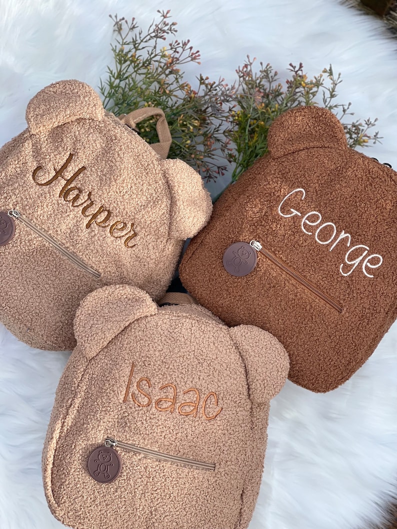 K&ouml;nnte beinhalten: Drei braune Teddyb&auml;r-Rucks&auml;cke mit personalisierten Namen: Harper, George und Isaac. Jeder Rucksack hat einen kleinen braunen B&auml;ren-Anh&auml;nger am Rei&szlig;verschluss.
