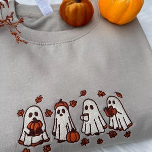 May include: A beige sweatshirt with an embroidered design of four cartoon ghosts with pumpkins and autumn leaves. Two small orange pumpkins sit on top of the sweatshirt. The design is in white, orange, and brown.