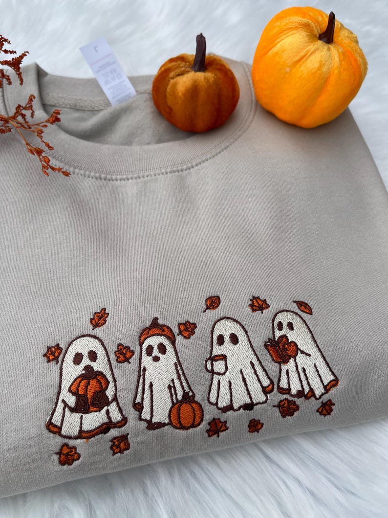 May include: A beige sweatshirt with an embroidered design of four cartoon ghosts with pumpkins and autumn leaves. Two small orange pumpkins sit on top of the sweatshirt. The design is in white, orange, and brown.